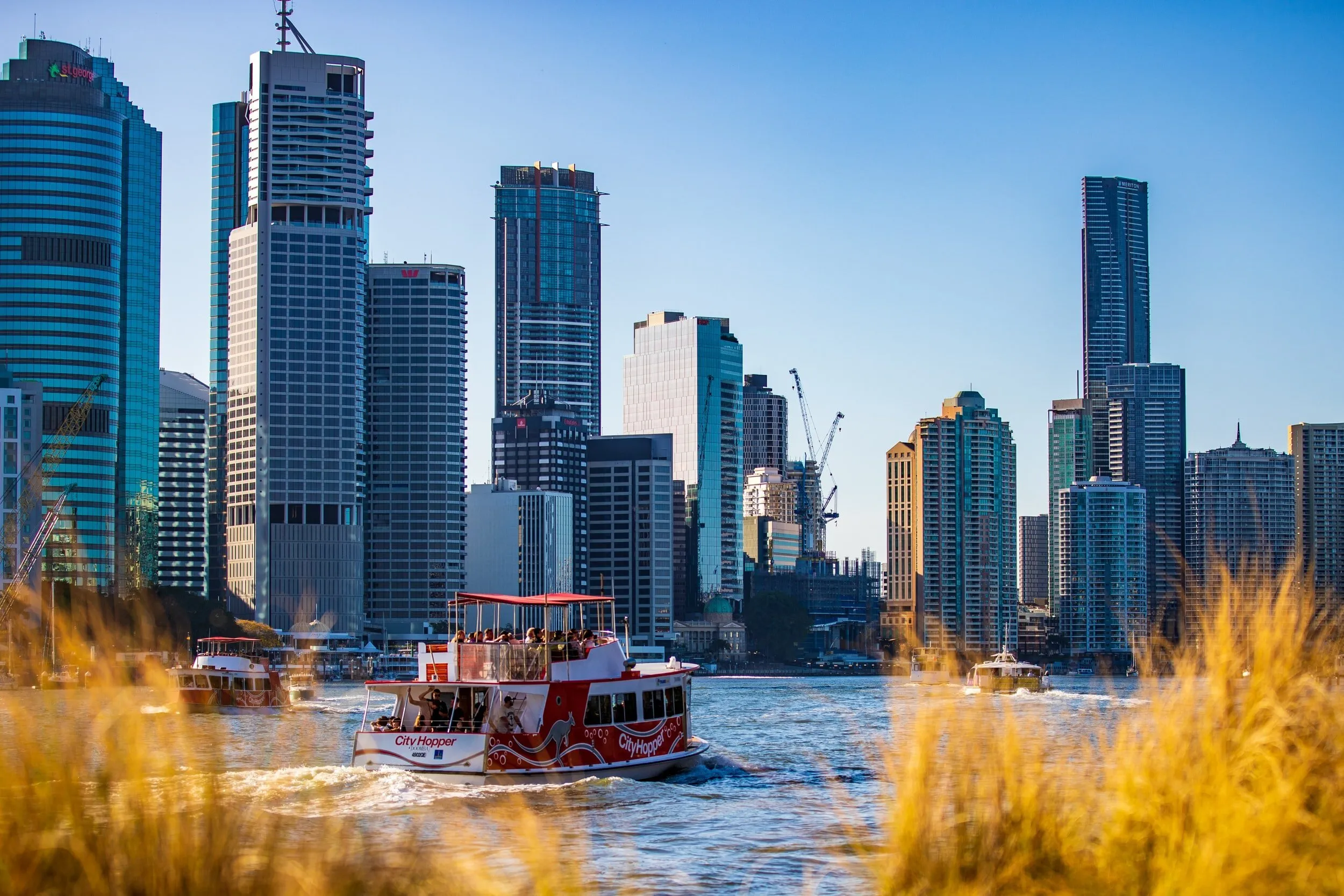 Scenic Brisbane cityscape from the river with a City Hopper ferry and modern skyscrapers under a blue sky, perfect for medical travel.