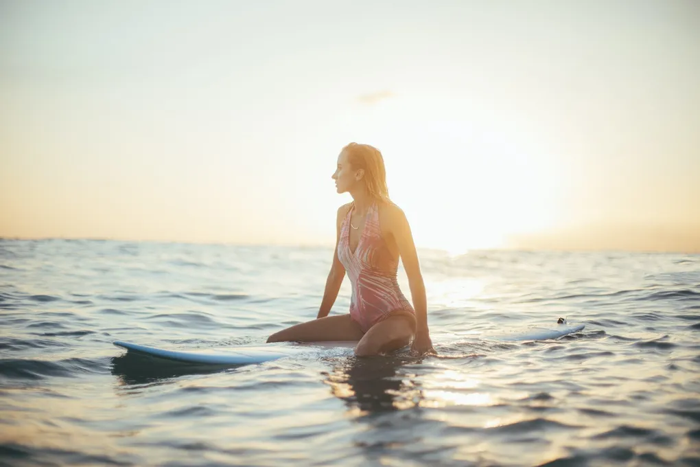 A young woman with wet hair sits on a surfboard in the calm ocean at sunset, looking towards the bright horizon.