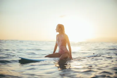 A young woman with wet hair sits on a surfboard in the calm ocean at sunset, looking towards the bright horizon.