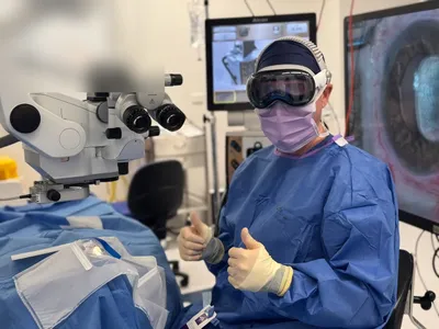 Dr David Gunn in surgical gown and Apple Vision Pro headset giving two thumbs up in a Brisbane operating theatre, with the Zeiss microscope and Alcon NGENUITY 3D displays behind him.