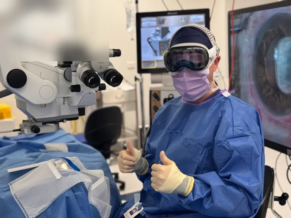 Dr David Gunn in surgical gown and Apple Vision Pro headset giving two thumbs up in a Brisbane operating theatre, with the Zeiss microscope and Alcon NGENUITY 3D displays behind him.