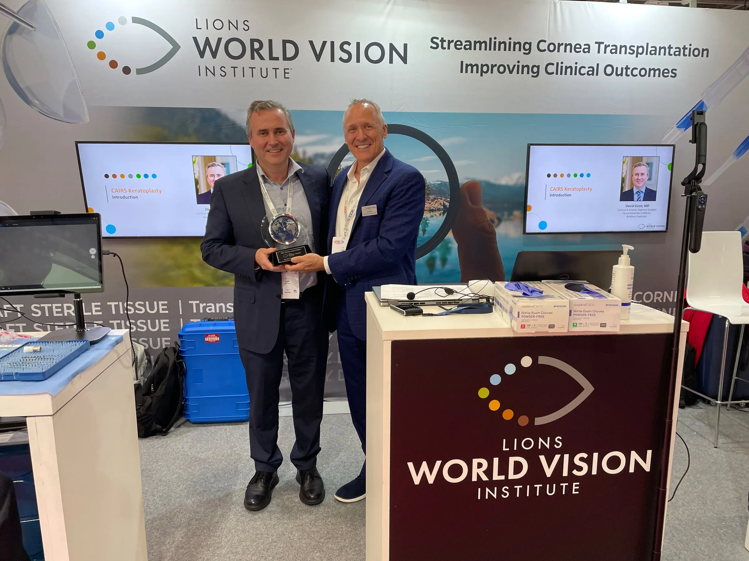 Dr David Gunn holding the glass clock presented by the Lions World Vision Institute at its stand at ESCRS 2026, beside an LWVI representative.