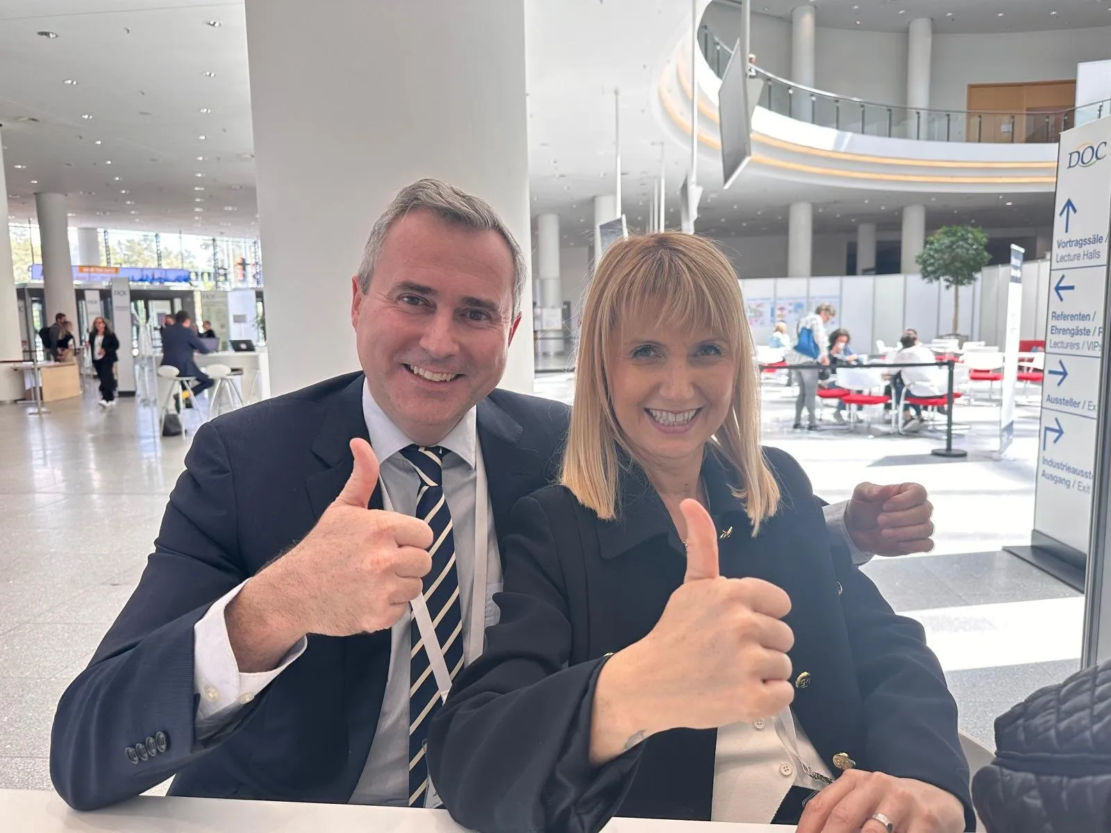 Dr. Gunn and colleague smiling, giving thumbs up at the German Ophthalmic Society (DOC) meeting. Conference hall background.