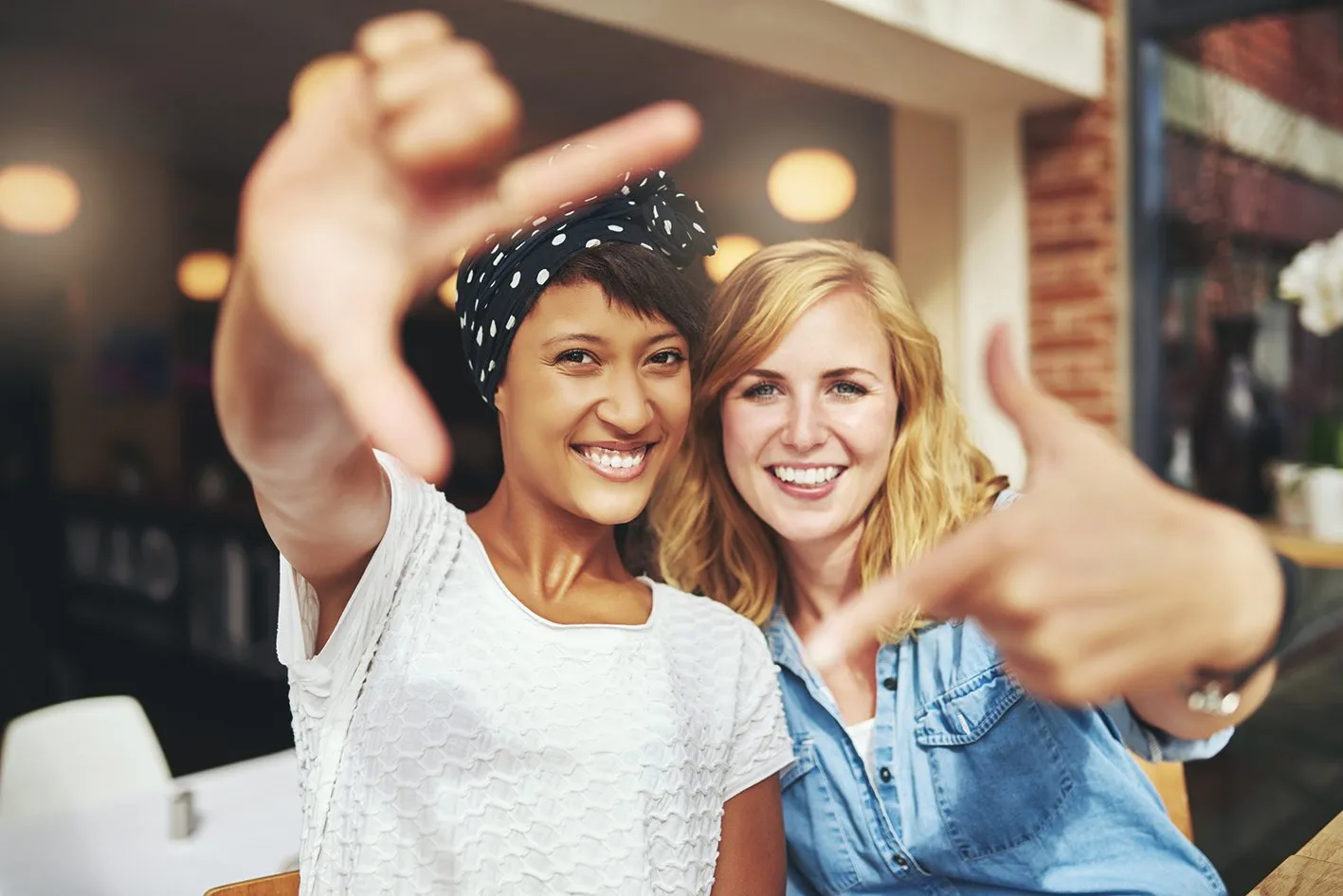 Two happy women, one blonde and one in a polka dot headscarf, smile and form hand frames, representing excellent vision.