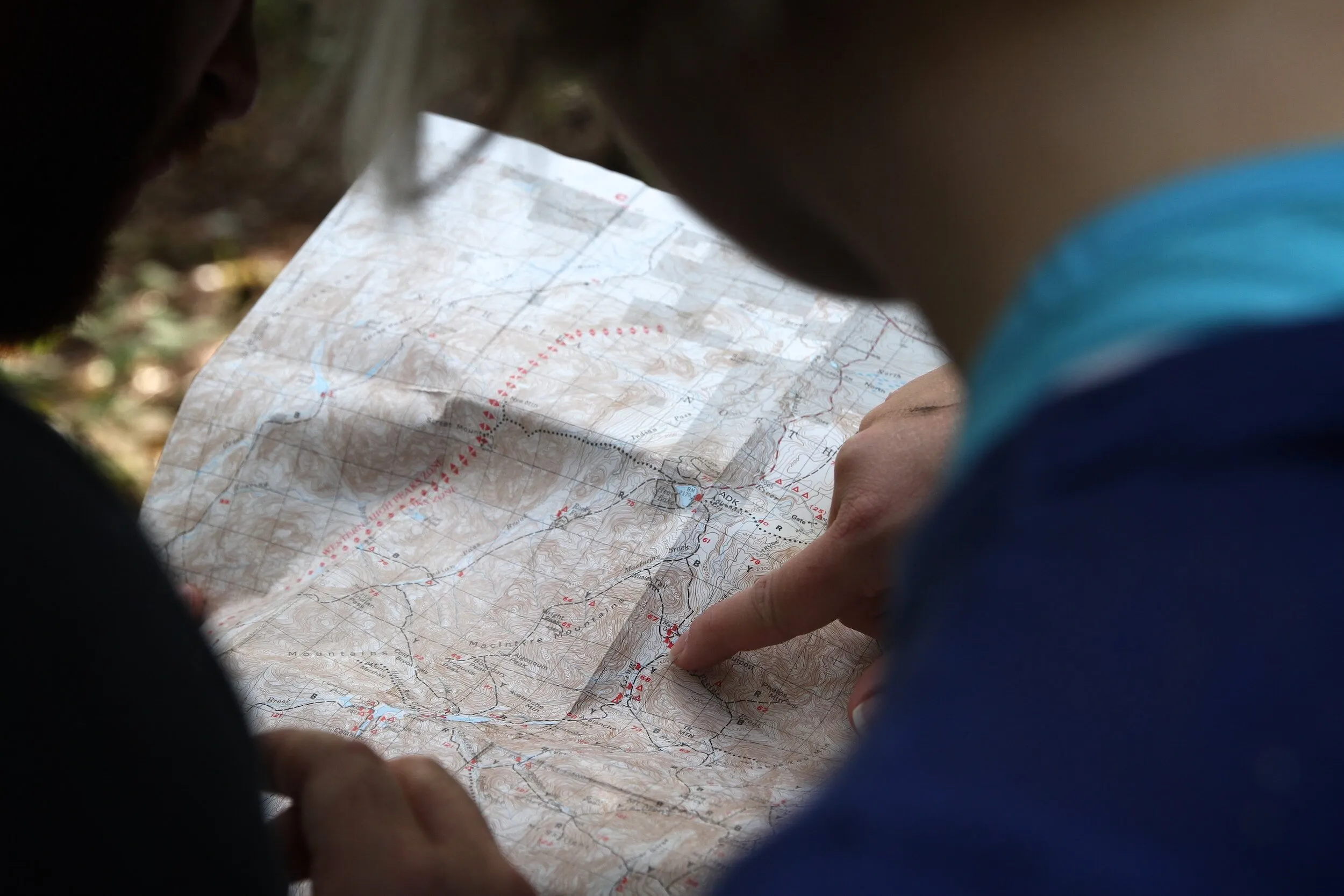 Close-up of two people, seen from behind, examining a topographic map. A finger points to a specific trail marker.