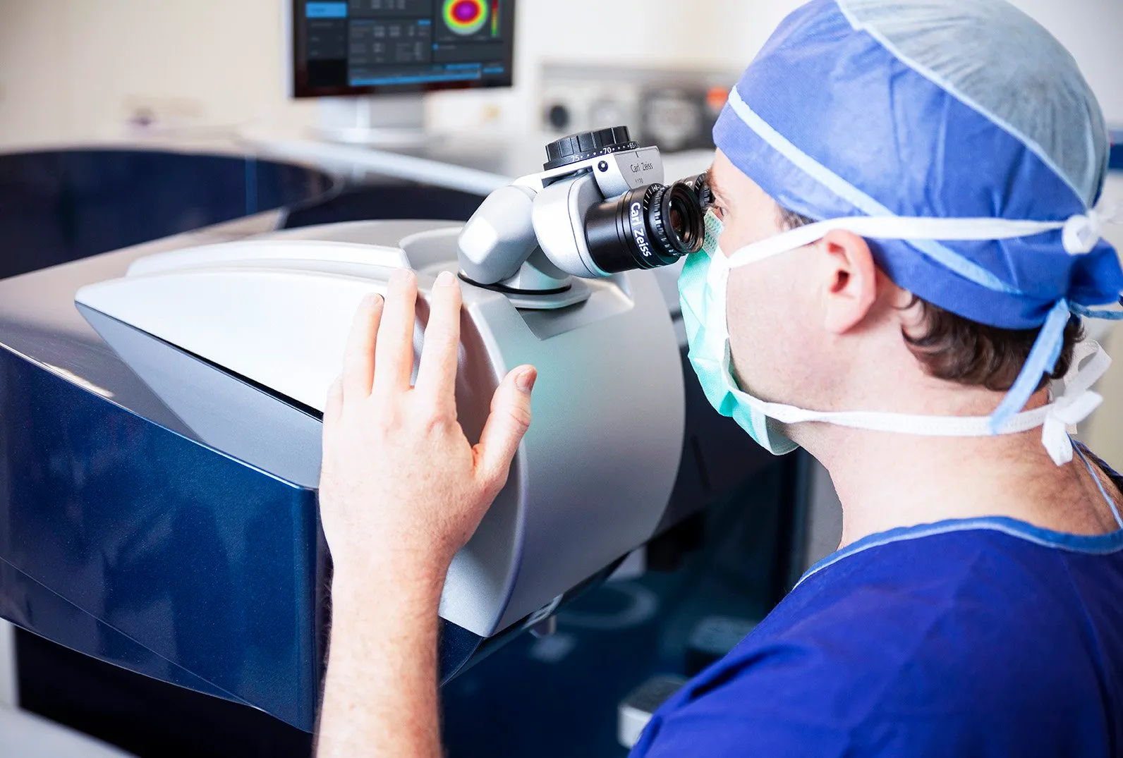 Ophthalmologist in scrubs and mask examining an eye with a Carl Zeiss ophthalmic machine; a diagnostic screen is visible.