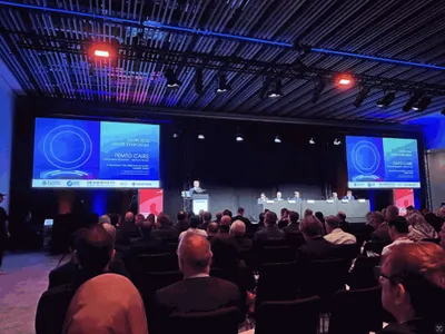Dr David Gunn at the lectern of the Ziemer satellite symposium at ExCeL London, ESCRS 2026, with the Femto CAIRS: Customised Segments title slide on both screens and the panel seated to his left.